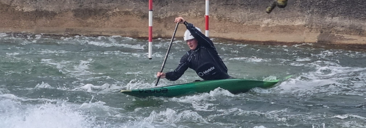 Aimee Collins in a green canoe on the water. Aimee is wearing a dark wetsuit and a white helmet.