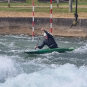 Aimee Collins in a green canoe on the water. Aimee is wearing a dark wetsuit and a white helmet.